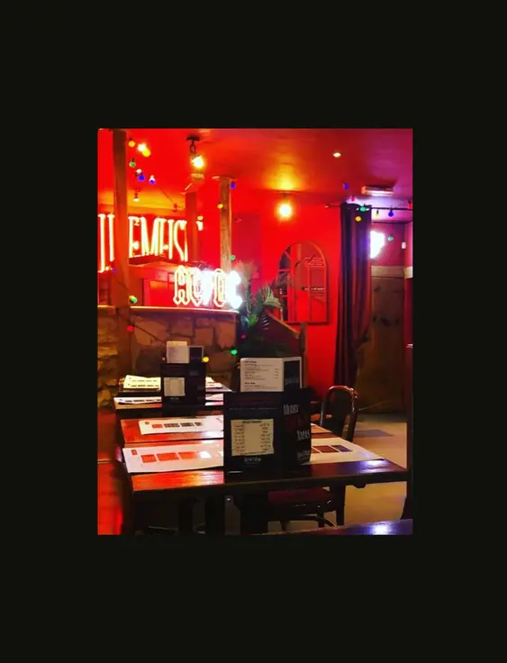 Vibrant bar interior with red lighting, neon signs, and colourful hanging bulbs over wooden tables.