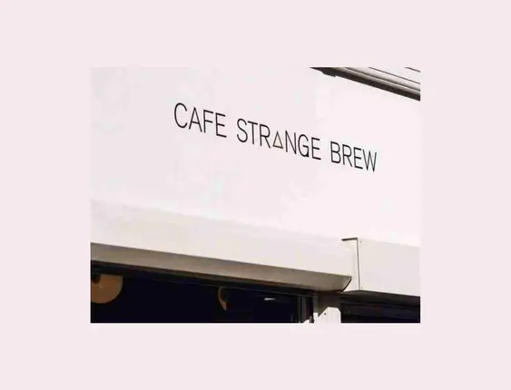 Outside Cafe Strange Brew, people wait in line under a sign. Sunlit, casual scene with plants at the cafe entrance.