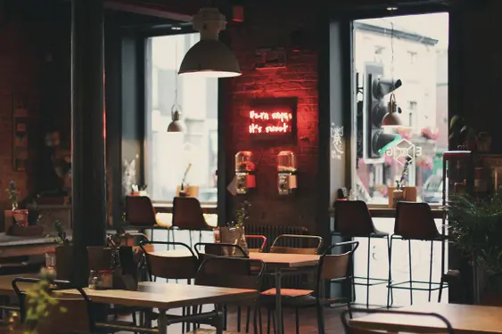 Dimly lit café with exposed brick walls and industrial-style lighting. Neon sign reads "Ezra says it's sweet" amidst modern furniture and large windows.