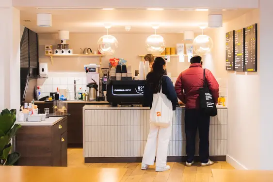 Customers order at a modern café counter, featuring a shiny espresso machine and menu boards. Overhead, large spherical lamps illuminate the warmly lit interior.