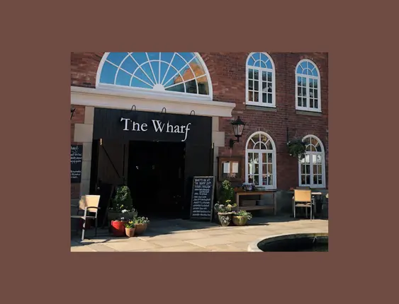 Large arched window tops the entrance of "The Wharf," a brick building with inviting outdoor seating under sunny skies.