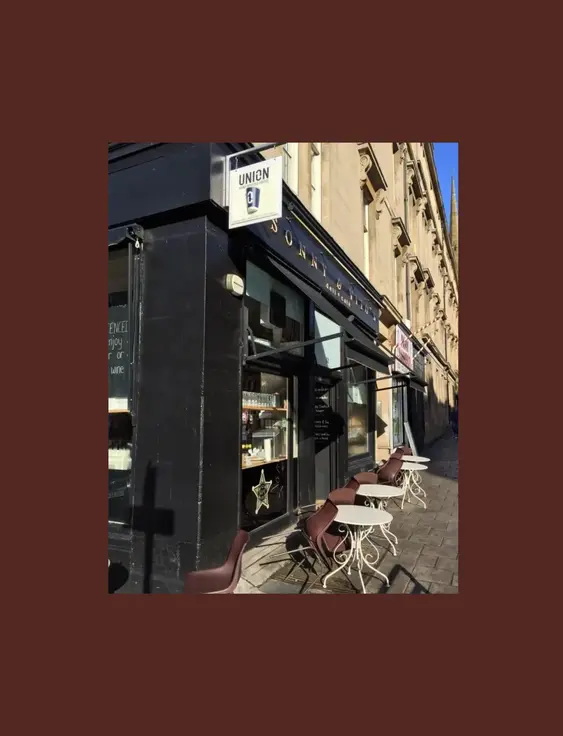 Exterior of a cafe on a corner, featuring outdoor seating with round tables, brown chairs, and a black facade under a sunny sky.