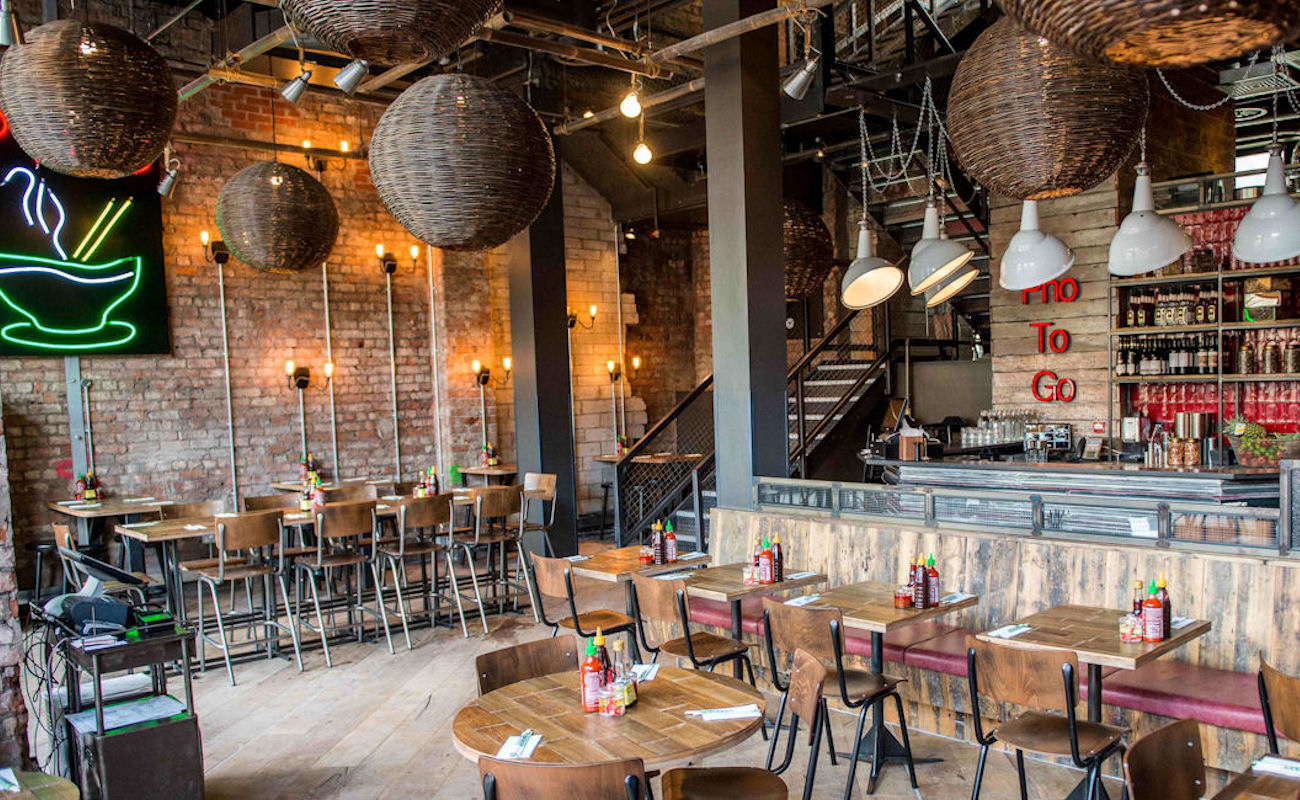 Spacious restaurant with exposed brick walls, large rattan pendant lights, and a neon "Pho to Go" sign above the bar. Wooden tables and chairs fill the dining area.