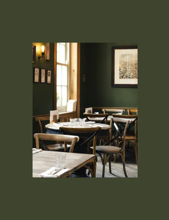 Intimate pub setting with dark green walls and wooden furniture. Natural light highlights the classic bentwood chairs and rustic tables set for dining. The walls are adorned with framed pictures and a large vintage-style painting.