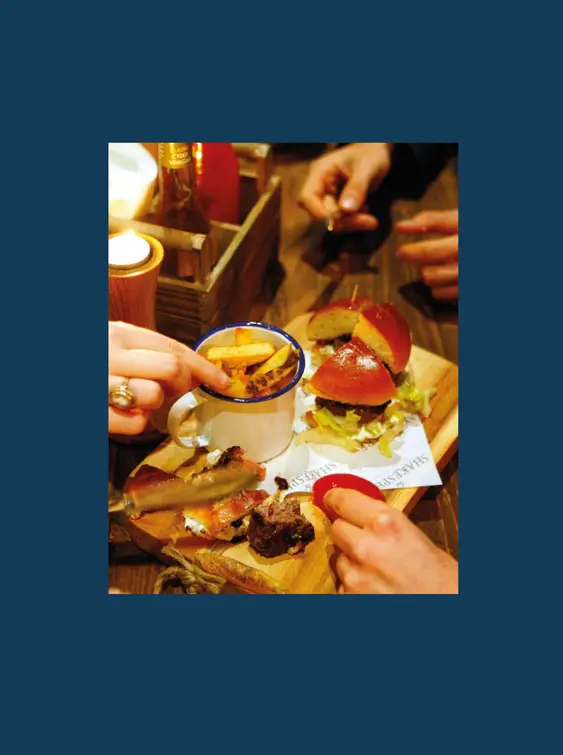 Four people's hands reaching towards a wooden board with a burger and chips on top of it.