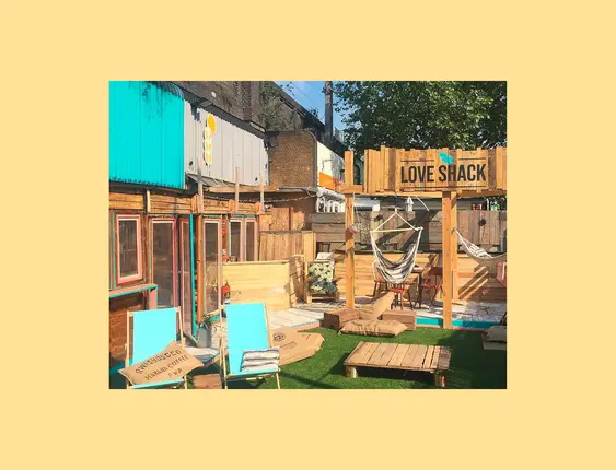 An outdoor restaurant space that shows multiple wooden decks and chairs for sitting. There is a small patch of astro turf at the front, upon which sits a blue deckchair.