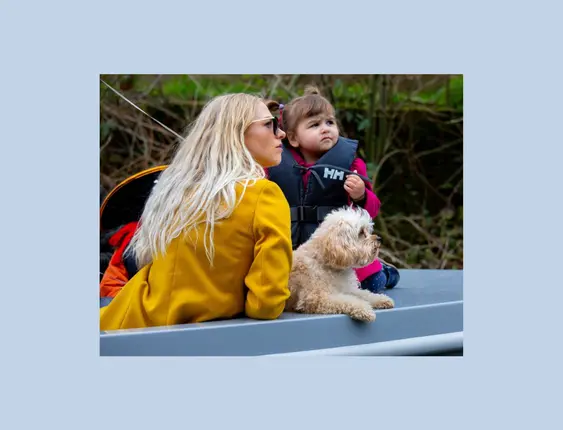 Women in yellow coat and sunglasses, child in safety jacket, and blond curly haired dog, sitting in a blue boat.