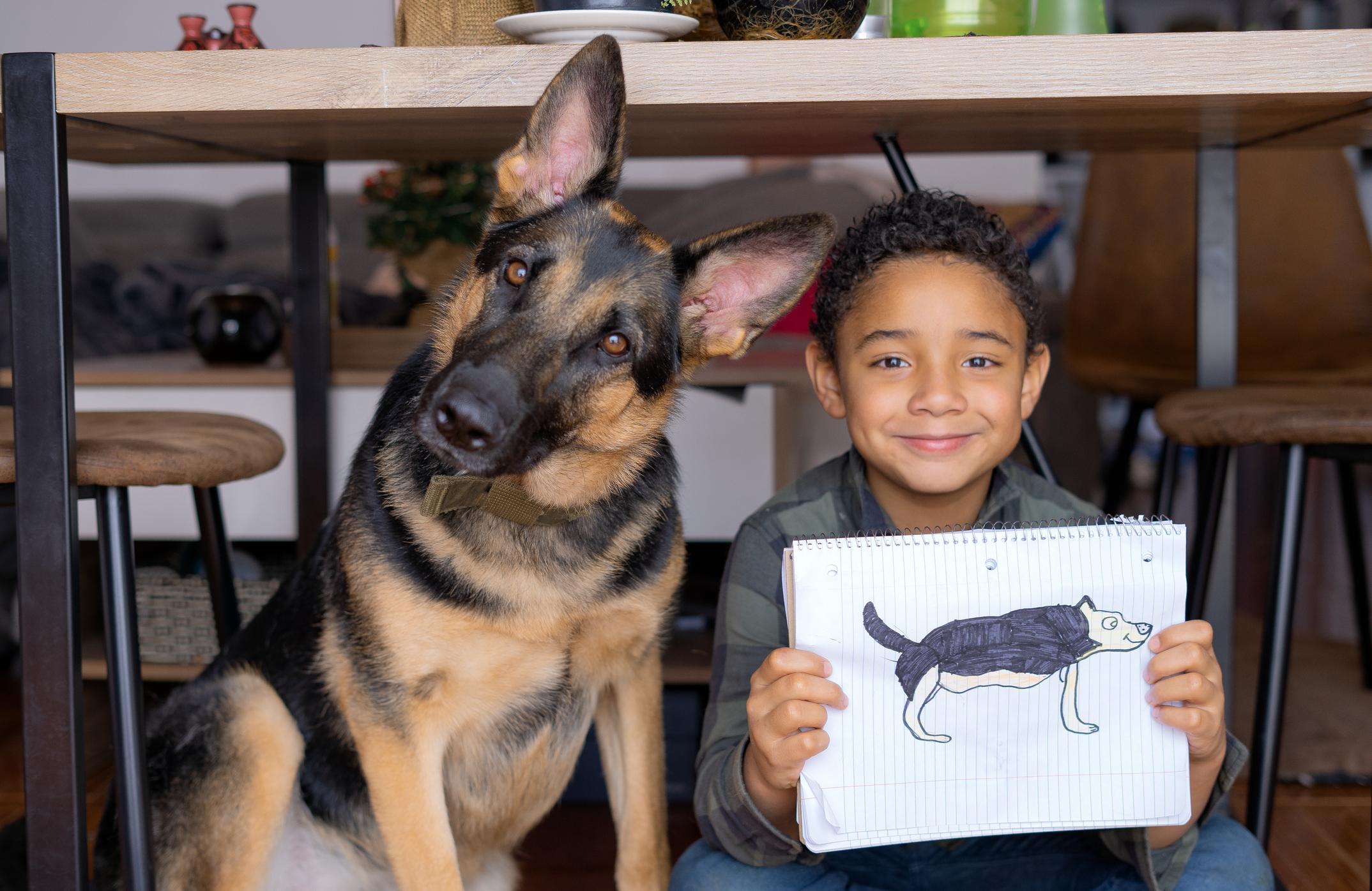 Portrait of a young black boy holding the drawing of his German Shepherd dog, which is sitting next to him. They are at home under the dining room