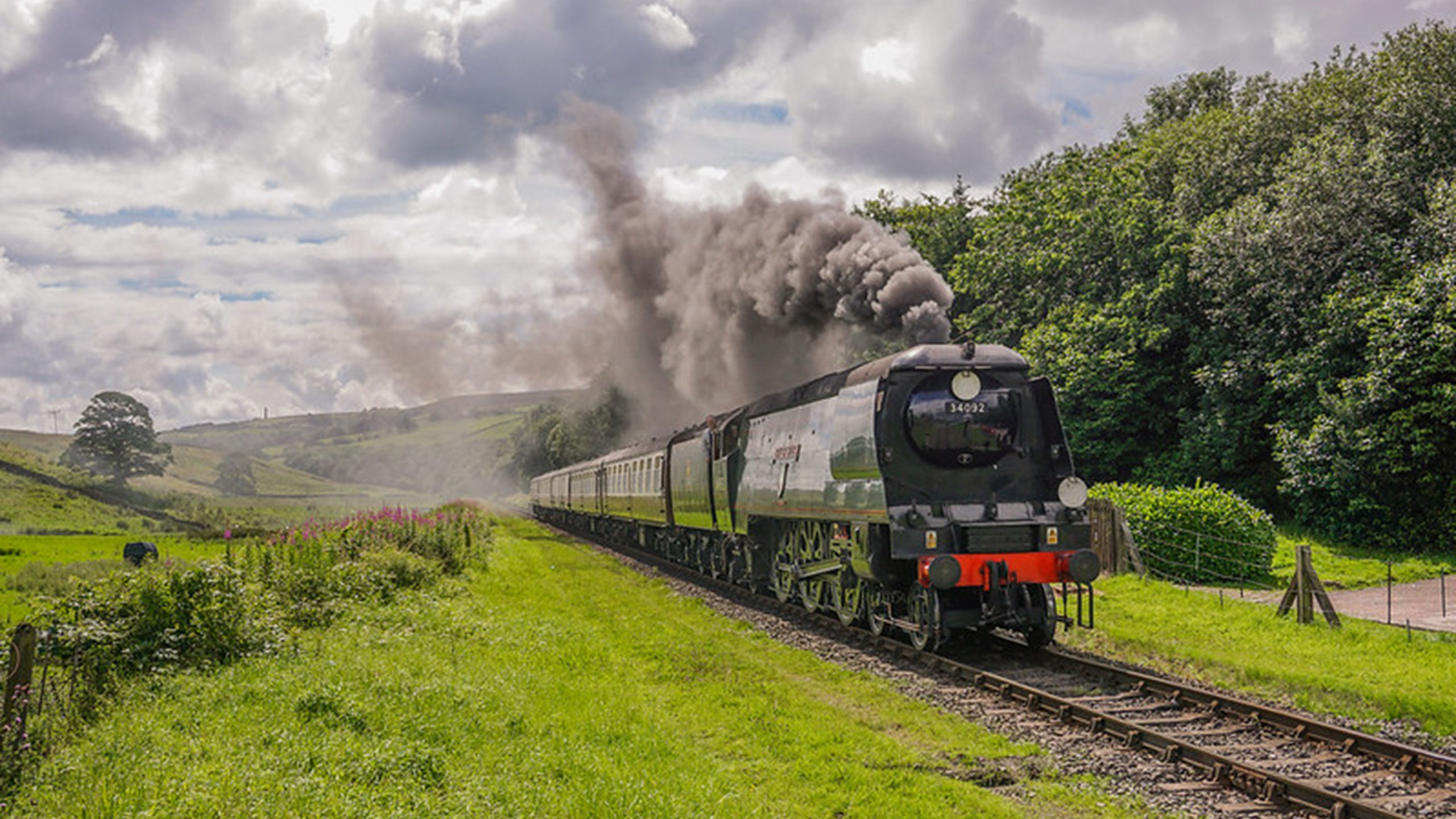 A vintage steam train billows thick smoke while traversing the lush green countryside under a partly cloudy sky, with hills in the backdrop and vibrant foliage alongside the tracks.